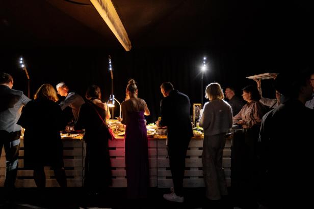 Mehrere Menschen stehen an einem beleuchteten Buffet und bedienen sich an Speisen in dunkler Umgebung.