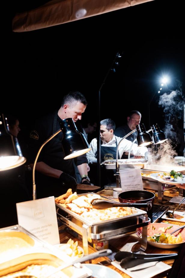 Mehrere Köche servieren unter warmem Licht verschiedene warme Speisen an einem Buffet mit dampfenden Gerichten.