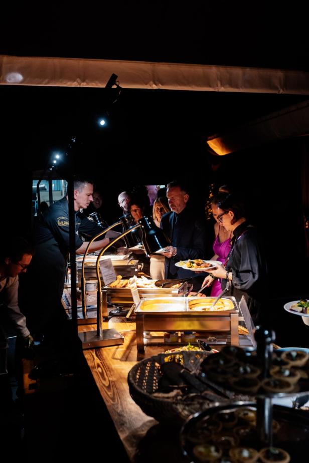 Mehrere Personen stehen an einem beleuchteten Buffet und lassen sich von einem Koch Speisen auf ihre Teller geben.