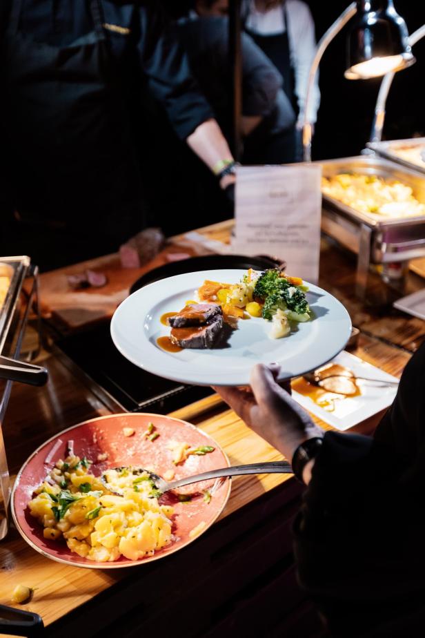Eine Person hält einen Teller mit Fleisch, Soße, Kartoffeln und Gemüse an einem Buffet mit verschiedenen Speisen.