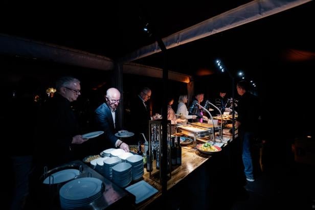 Mehrere Personen bedienen sich an einem beleuchteten Buffet mit verschiedenen Speisen in dunkler Umgebung.