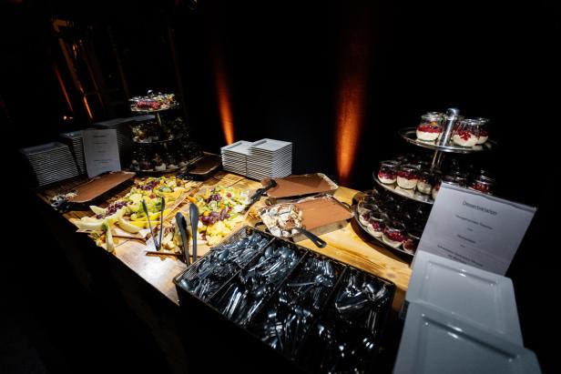 Ein Buffet mit verschiedenen Desserts, frischem Obst, Tiramisu, Besteck und gestapelten Tellern auf einem Holztisch.