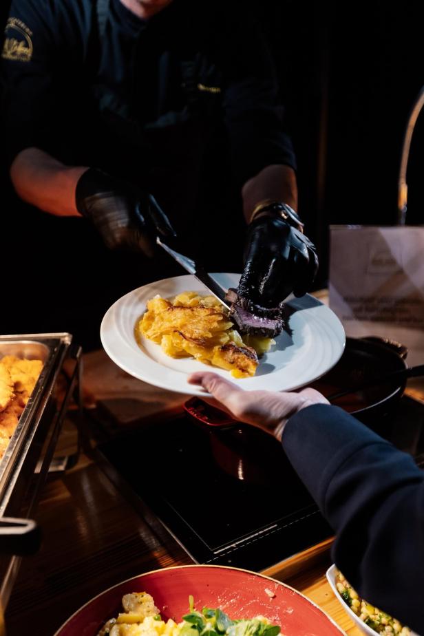 Ein Koch mit schwarzen Handschuhen serviert einem Gast Kartoffelgratin und Fleisch auf einem weißen Teller.