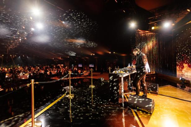 Ein DJ steht an einem Mischpult auf einer Bühne, während viele Gäste an festlich gedeckten Tischen sitzen und Lichteffekte den Raum erhellen.
