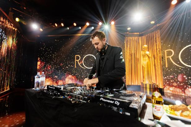 Ein DJ in schwarzer Kleidung legt an einem Mischpult auf einer festlich beleuchteten Bühne mit goldener Statue auf.