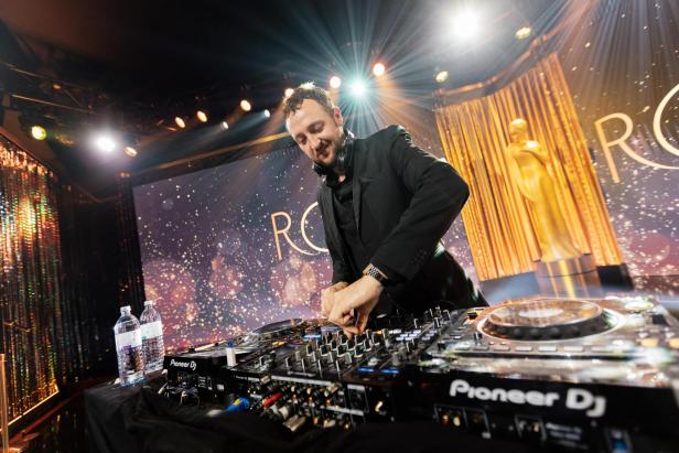 Ein DJ in schwarzem Anzug legt an einem Mischpult auf einer festlich beleuchteten Bühne mit goldener Statue auf.