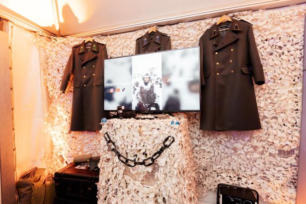 Drei Militärmäntel hängen an einer Wand mit Tarnnetz, darunter steht ein Monitor mit Social-Media-Hinweisen und Ausrüstung.