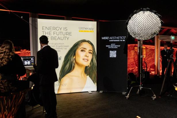 Zwei Personen stehen vor einem großen Werbeplakat mit einer Frau und einem Laptop, daneben ein Fotolicht und ein schwarzes Werbeschild.