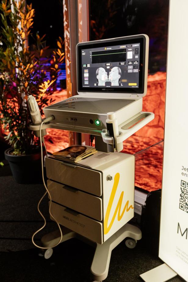 Ein medizinisches Gerät mit Bildschirm und Handstück steht auf Rollen neben einem Fenster, auf dem Monitor ist ein Gesicht abgebildet.