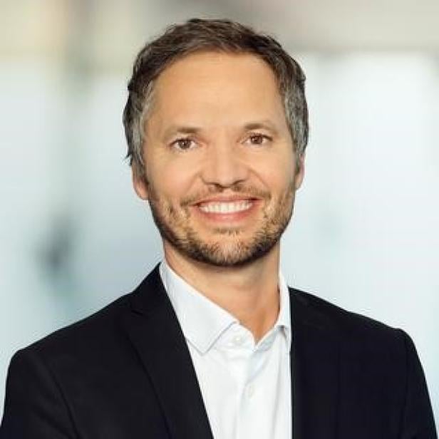 Mann im Anzug mit weißem Hemd lächelt vor unscharfem Hintergrund.