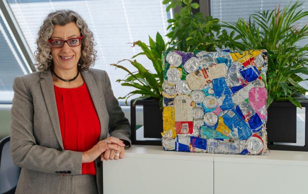 Eine Frau mit lockigen Haaren steht neben einem Kunstwerk aus gepressten Getränkedosen und grünen Pflanzen.