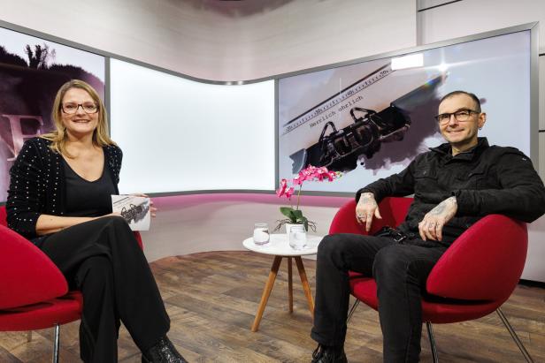 Zwei Personen sitzen in roten Sesseln in einem modernen Studio, zwischen ihnen steht ein kleiner Tisch mit Blumen und Wassergläsern.