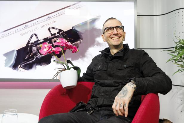 Ein lächelnder Mann mit Brille sitzt in einem roten Sessel, hält eine Orchidee und im Hintergrund ist eine Schreibmaschine zu sehen.