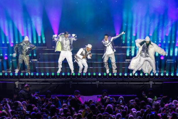 Fünf Männer in silberner Kleidung tanzen auf einer Bühne mit bunten Lichtern vor einem begeisterten Publikum.