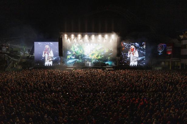 Eine große Menschenmenge steht vor einer beleuchteten Konzertbühne mit zwei großen Leinwänden und tropischem Bühnenbild.