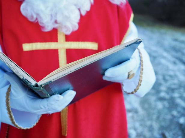 Eine Person mit weißem Bart und roten Gewand mit Kreuz hält ein aufgeschlagenes Buch in behandschuhten Händen.