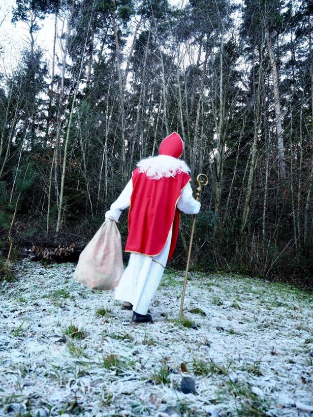 Nikolaus mit Sack und Stab wandert durch einen winterlichen Wald.