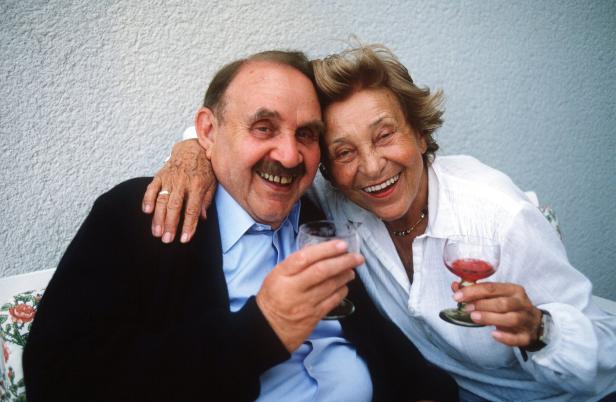 Ein älteres Paar sitzt eng umarmt zusammen und lächelt fröhlich, während beide ein Glas Wein halten.