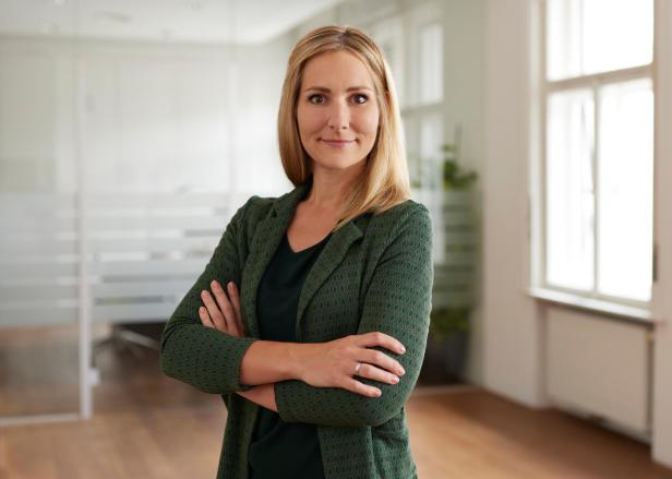 Eine Frau mit blondem Haar steht mit verschränkten Armen in einem modernen, hellen Büro und lächelt leicht.