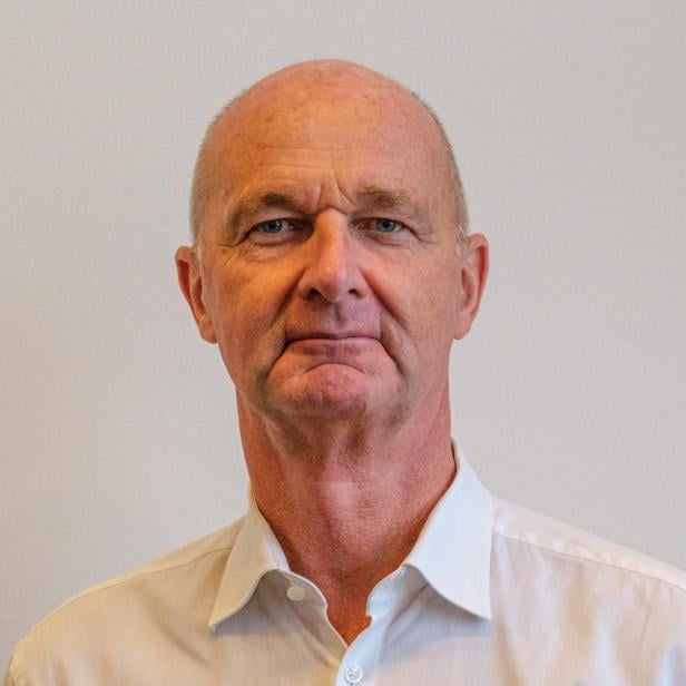 Ein älterer Mann mit Glatze und weißem Hemd blickt ernst in die Kamera vor hellem Hintergrund.