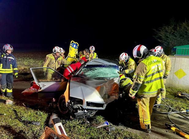 Mehrere Feuerwehrleute stehen um einen demolierten, grauen Pkw herum.