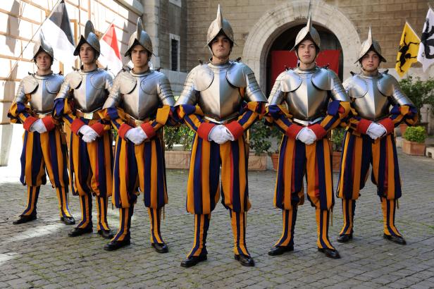 Sechs Gardisten in bunter Uniform und Rüstung stehen nebeneinander vor einem historischen Gebäude.