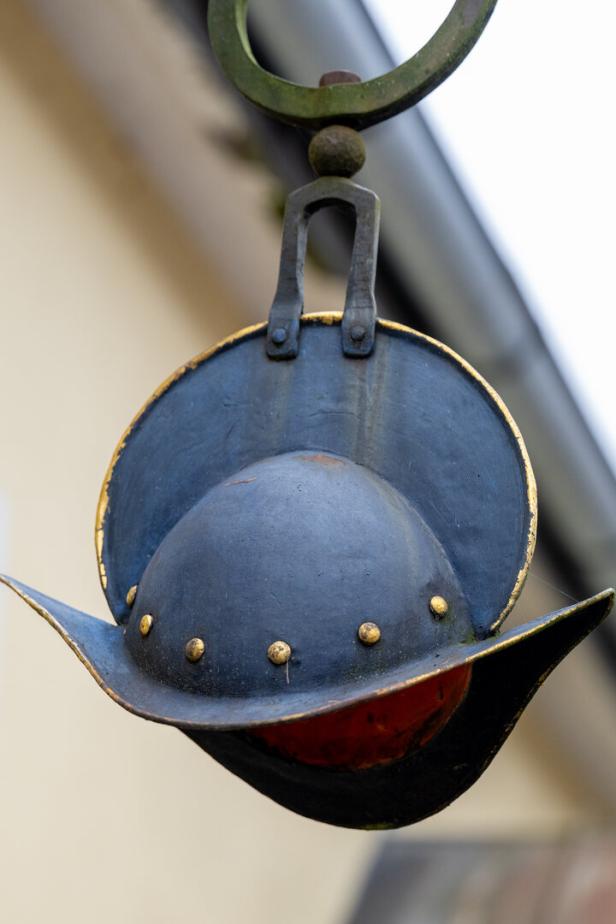 Ein schwarzer, metallener Hut mit goldenen Nieten und breiter Krempe hängt an einer Halterung.