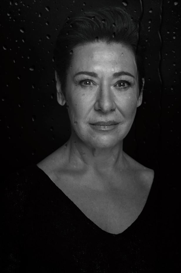 Eine Frau mit kurzem Haar blickt ernst in die Kamera, im Hintergrund sind Wassertropfen auf einer Scheibe zu sehen.