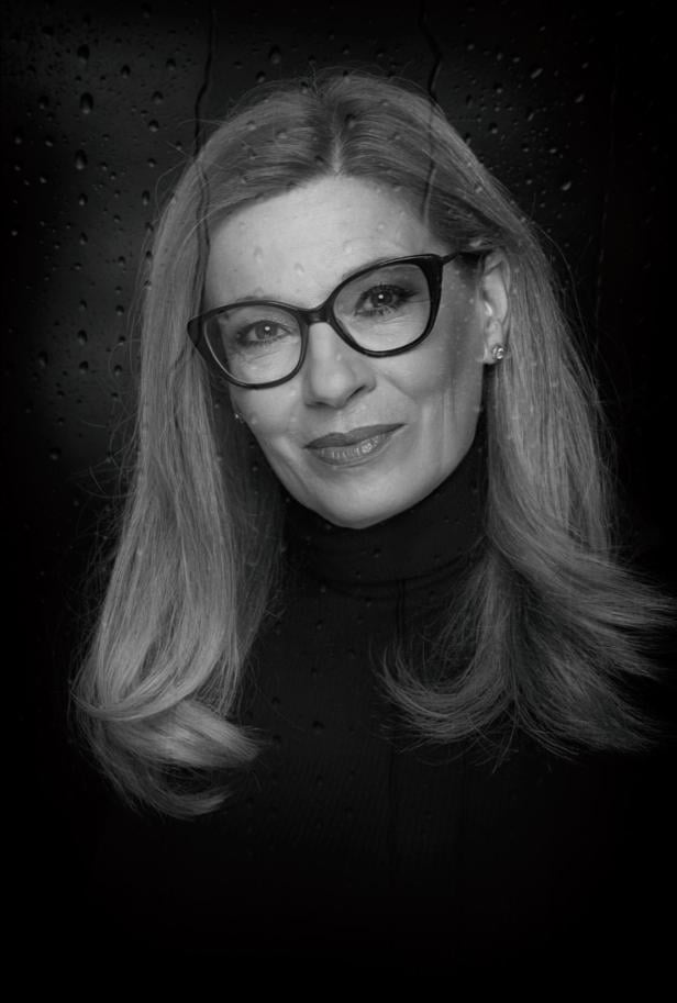 Eine Frau mit kurzem Haar blickt ernst in die Kamera, im Hintergrund sind Wassertropfen auf einer Scheibe zu sehen.