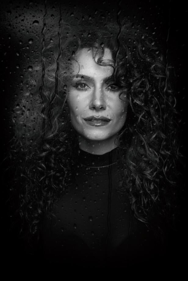 Eine Frau mit kurzem Haar blickt ernst in die Kamera, im Hintergrund sind Wassertropfen auf einer Scheibe zu sehen.