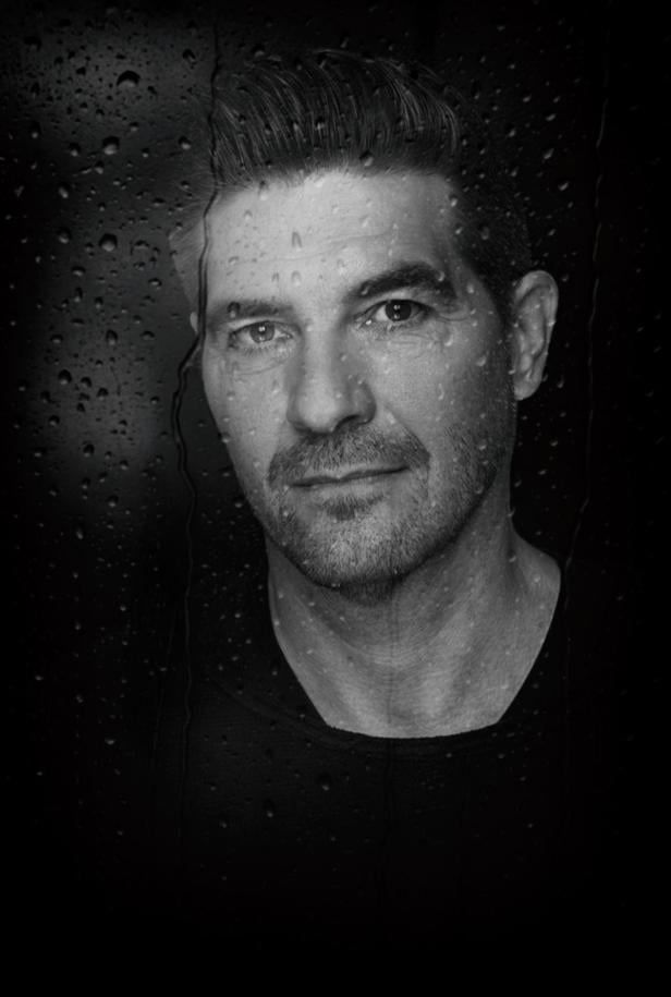 Eine Frau mit kurzem Haar blickt ernst in die Kamera, im Hintergrund sind Wassertropfen auf einer Scheibe zu sehen.