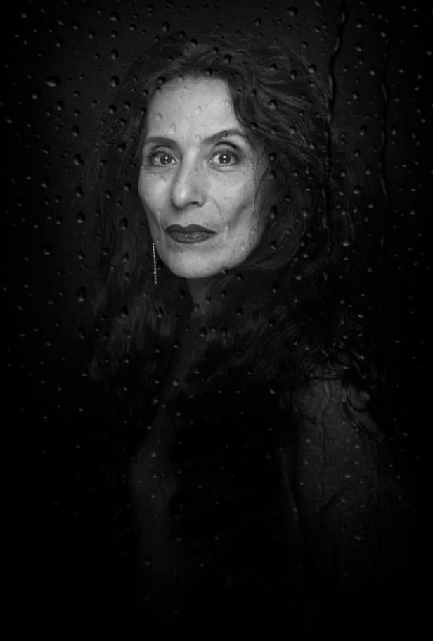 Eine Frau mit kurzem Haar blickt ernst in die Kamera, im Hintergrund sind Wassertropfen auf einer Scheibe zu sehen.