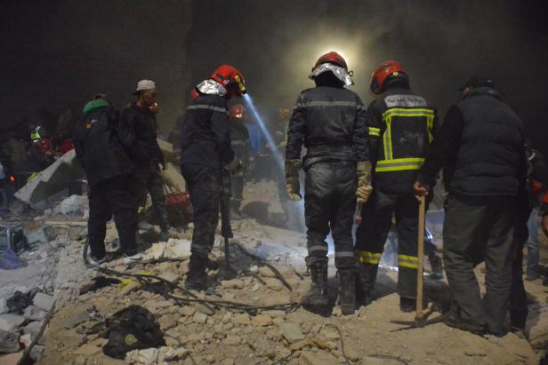 Mehrere Rettungskräfte mit Helmen und Werkzeugen suchen nachts in Trümmern nach Überlebenden.