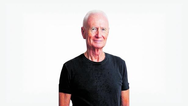 Ein älterer Mann mit kurzen weißen Haaren trägt ein schwarzes T-Shirt und lächelt leicht vor hellem Hintergrund.