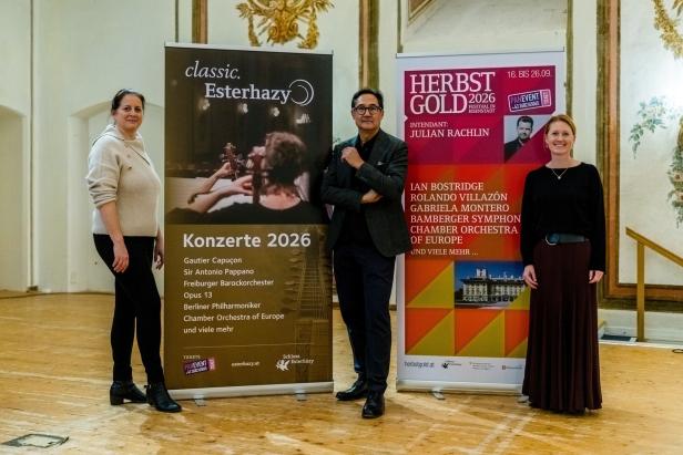 Drei Personen stehen in einem prunkvollen Saal neben zwei Werbebannern für klassische Konzerte und ein Musikfestival.