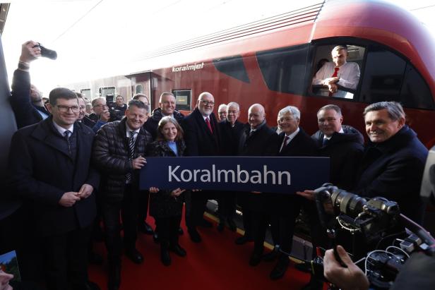 Eine Gruppe von Menschen hält ein Schild mit der Aufschrift "Koralmbahn" vor einem roten Zug.