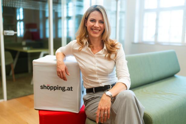 Eine Frau sitzt lächelnd auf einem Sofa und lehnt an einem Würfel mit der Aufschrift "shöpping.at"
