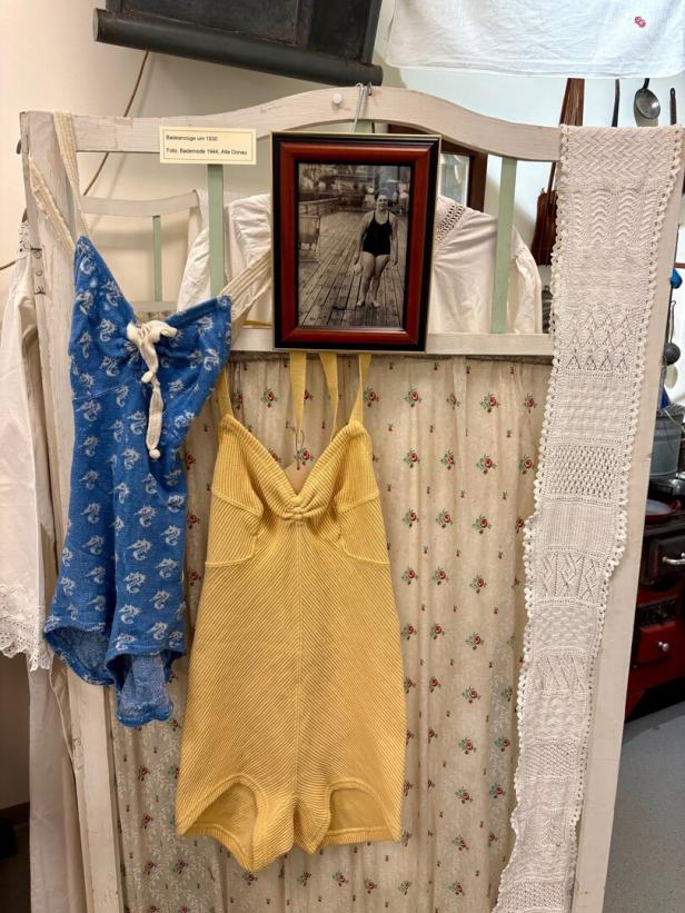 Zwei historische Badeanzüge, ein blauer und ein gelber, hängen neben einem gerahmten Schwarzweißfoto einer Frau im Badeanzug.