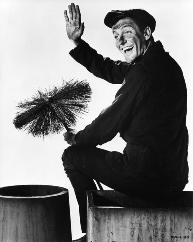 Ein lachender Schornsteinfeger in Arbeitskleidung sitzt auf einem Schornstein und hält eine Reinigungsbürste, während er winkt.