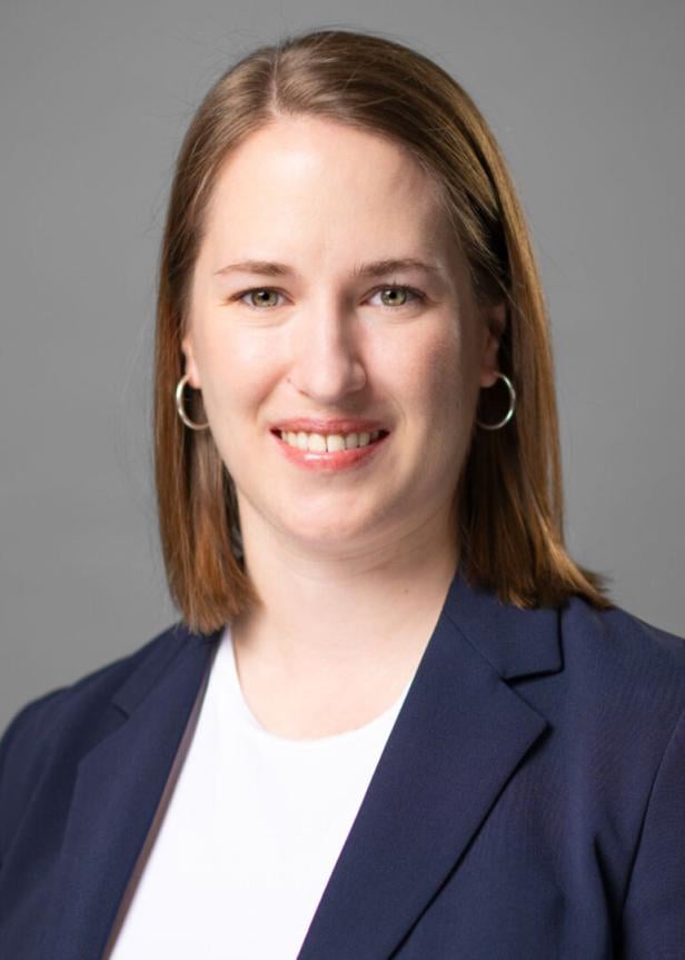 Junge Frau mit schulterlangen braunen Haaren, silbernen Ohrringen und dunkelblauem Blazer vor grauem Hintergrund.