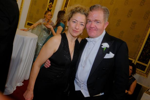 Ein Paar in eleganter Abendgarderobe steht eng beieinander, im Hintergrund unterhalten sich weitere festlich gekleidete Personen.