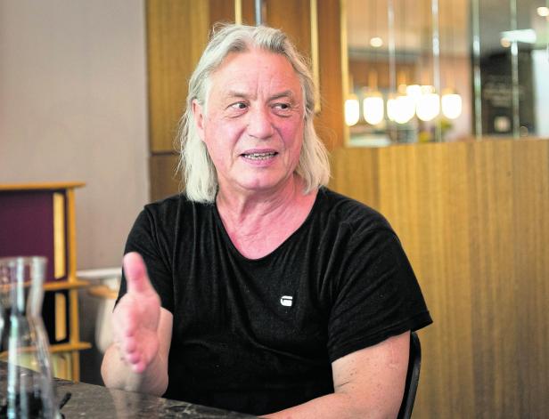 Ein älterer Mann mit langen grauen Haaren und schwarzem T-Shirt sitzt an einem Tisch und gestikuliert mit der rechten Hand.