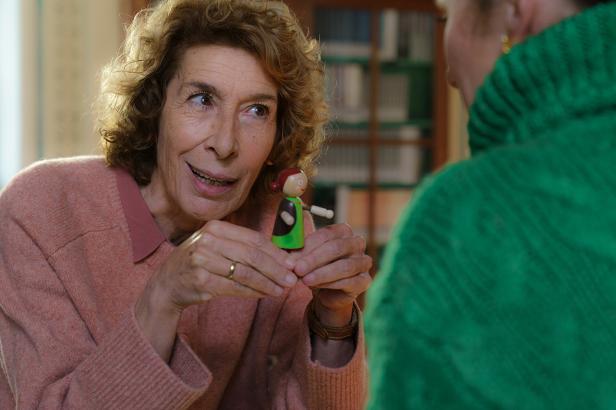 Eine ältere Frau im rosa Pullover hält eine kleine Holzpuppe in den Händen.