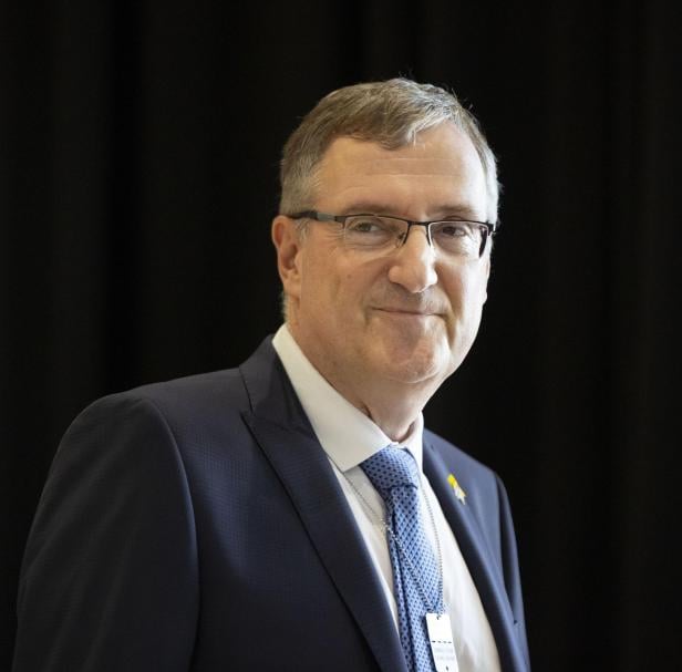 Ein Mann mit Brille und Anzug lächelt vor einem dunklen Hintergrund.