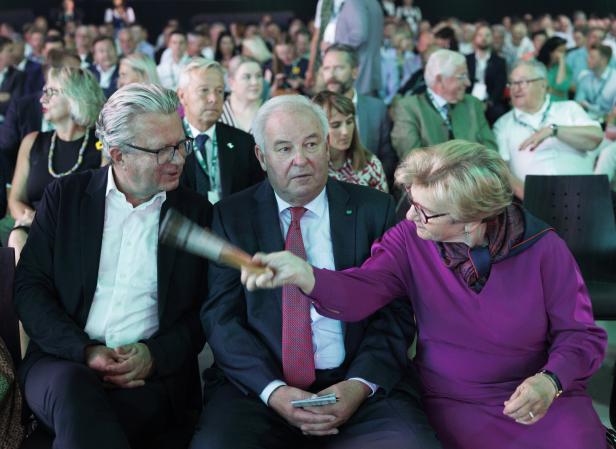 Eine Frau in lila Kleidung neben zwei Männnern in Anzügen, im Hintergrund viele weitere Menschen