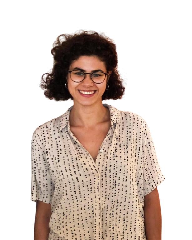 Junge Frau mit lockigen dunklen Haaren, Brille und gemusterter Bluse lächelt freundlich vor weißem Hintergrund.