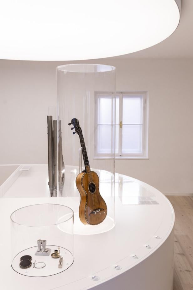 Eine Gitarre steht in einem durchsichtigen Zylinder auf einem weißen, runden Tisch in einem hellen Raum.