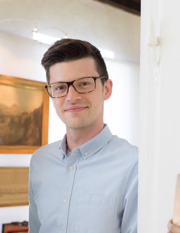 Ein junger Mann mit Brille und hellblauem Hemd steht lächelnd in einem hellen Raum mit Gemälden an der Wand.
