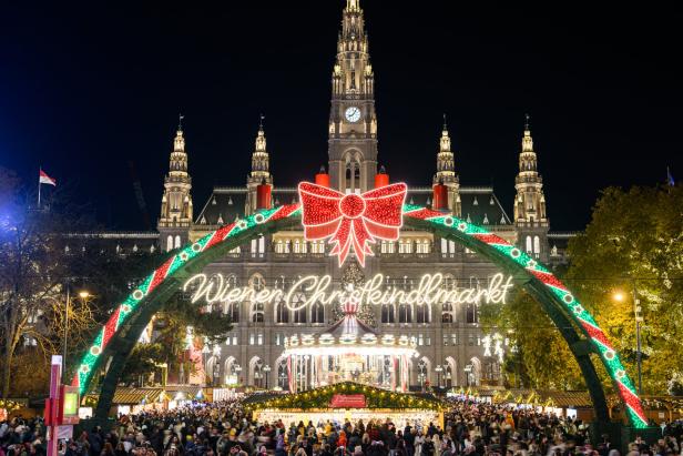 Festlich beleuchteter Weihnachtsmarkt mit Menschenmenge vor einem historischen Gebäude und großem Schriftzug „Wiener Christkindlmarkt“.