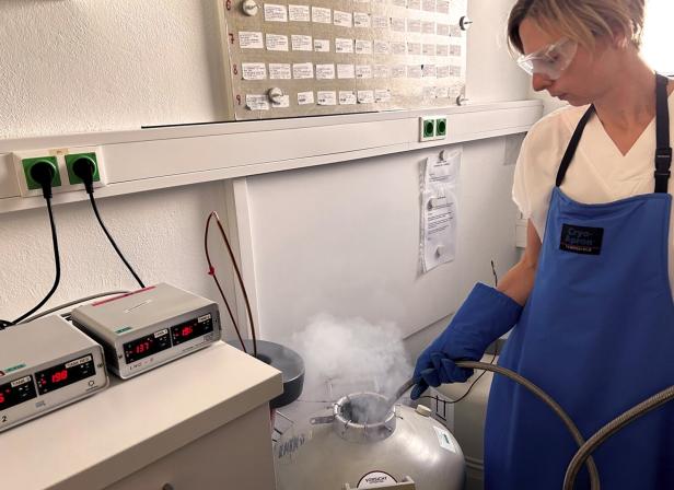 Eine Person mit Schutzbrille und blauer Schürze arbeitet an einem dampfenden Kryotank in einem Labor.